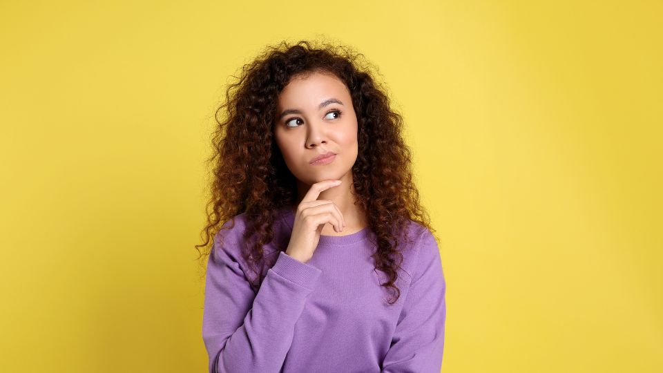 Femme réfléchissante sur fond jaune Une femme avec des cheveux bouclés portant un pull violet, réfléchissant avec une main sur le menton sur un fond jaune.