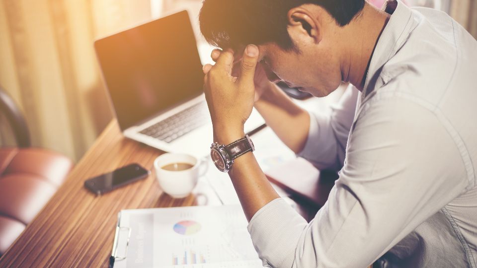 Stress lié aux finances personnelles Un homme assis à son bureau, tenant sa tête entre ses mains avec des graphiques financiers et un ordinateur portable à côté de lui.