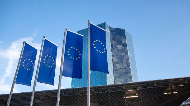 Drapeaux de l'Union Européenne devant un bâtiment moderne en verre Quatre drapeaux de l'Union Européenne flottant devant un bâtiment moderne en verre sous un ciel bleu clair.